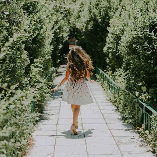 photography of woman in white and red floral midi dress walking on pathway